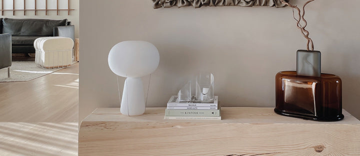 Japanda interior, wit a side table on which are presented the NUDE Blow lamp in opal white, a set of books on top of which are the Mist Lights votives in clear as well as the Layers vase wide in amber and Layers vase tall in grey with some brenches