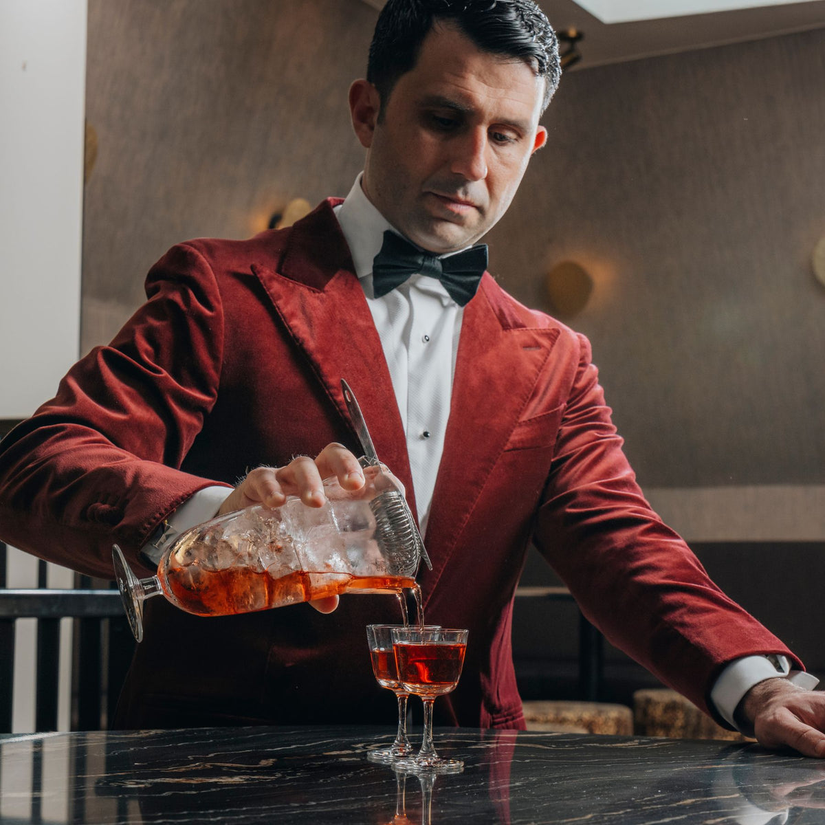 Giorgio BarGiani in a red velvet blazer, white dress shirt, and black bow tie pours an amber-colored cocktail from his NUDE doublepour mixing glass shaker into the new NUDE welcome drink glass on a dark marble table.