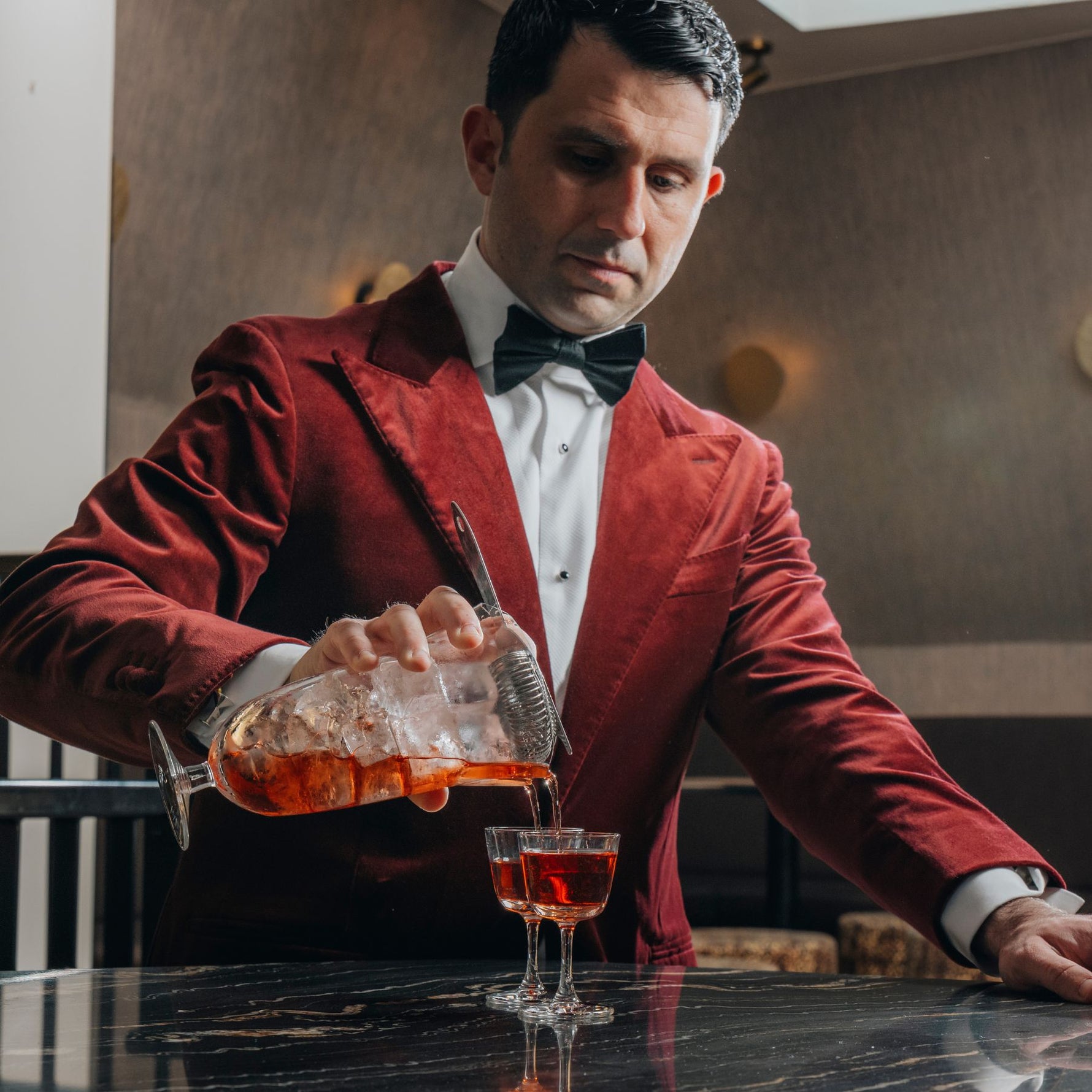 Giorgio BarGiani in a red velvet blazer, white dress shirt, and black bow tie pours an amber-colored cocktail from his NUDE doublepour mixing glass shaker into the new NUDE welcome drink glass on a dark marble table.