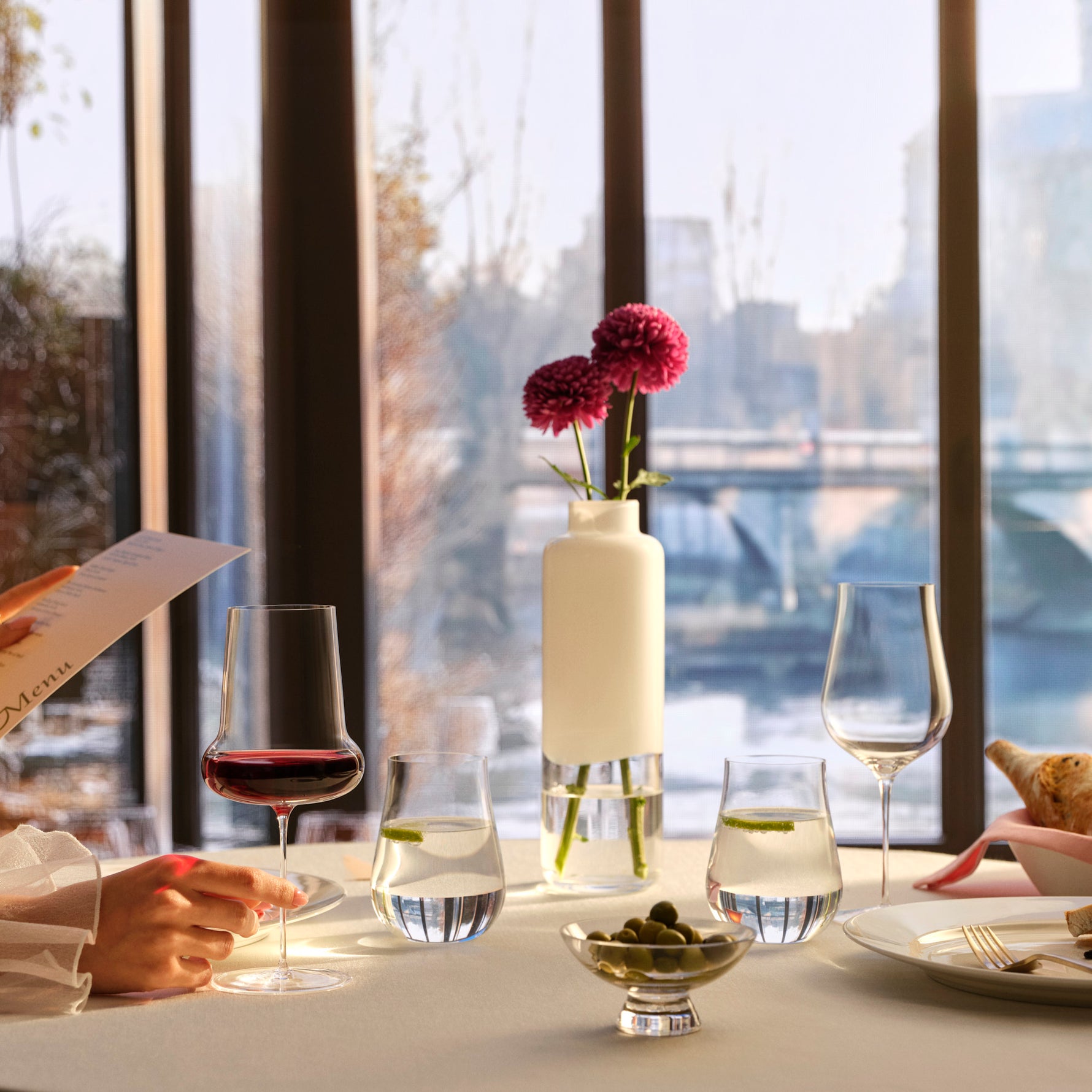 Dining table setting with the Nude glass Ghost Zero Belly Red wine glass and the Ghost Zero Tulip White wine glas, placed on the table with a Nude glass Magnolia vase of flowers in a restaurant, while also served with the Ghost Zero waterglasses and the Silhouette serving bowl. 