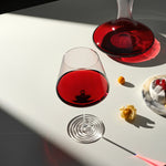 A clear stemmed wine glass and a matching decanter filled with deep red liquid, arranged on a white surface with sharp sunlight shadows and a small dessert plate nearby.