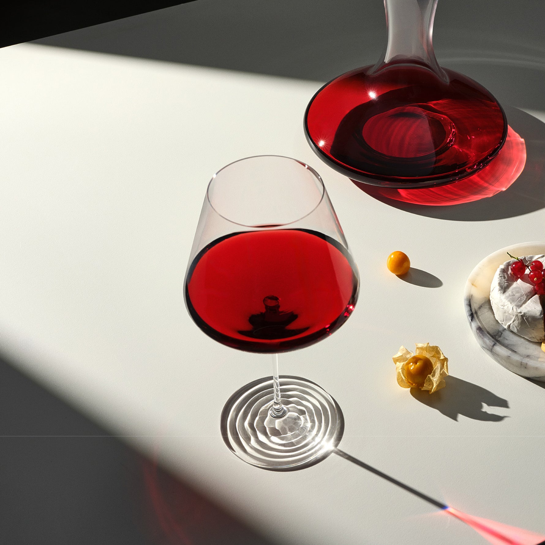 A clear stemmed wine glass and a matching decanter filled with deep red liquid, arranged on a white surface with sharp sunlight shadows and a small dessert plate nearby.