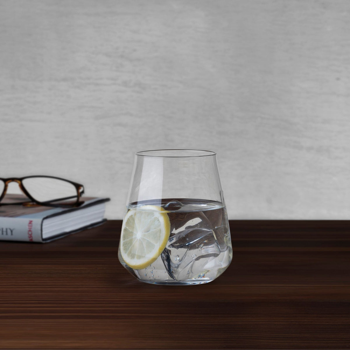 NUDE Salute tumbler with water, ice and lemon slice beside a book and reading glasses on a wooden table