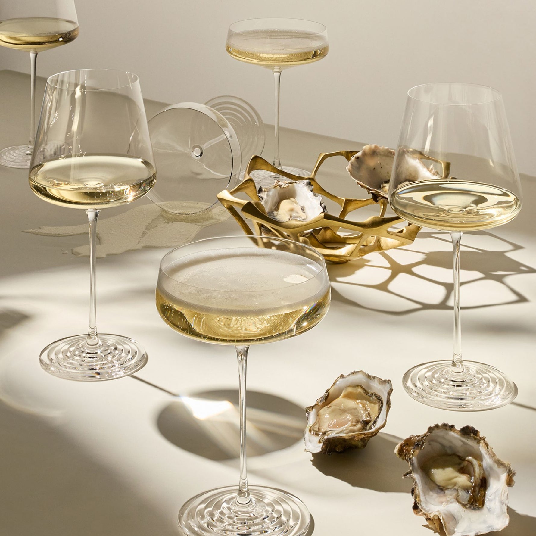 A set of elegant clear glass wine and cocktail glasses of the NUDE x Tom Dixon Splash collection arranged on a warm beige surface with strong sunlight casting shadows. The glasses include a large white wine glass, two coupe-style glasses, a lowball glass, and a gold geometric tray holding oyster shells.