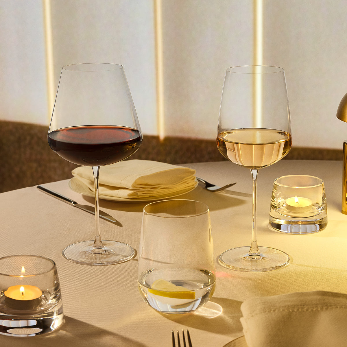 Two NUDE Sera wine glasses on a softly lit dining table, one filled with red wine and one with white wine, alongside candle holders, folded napkins, and cutlery.
