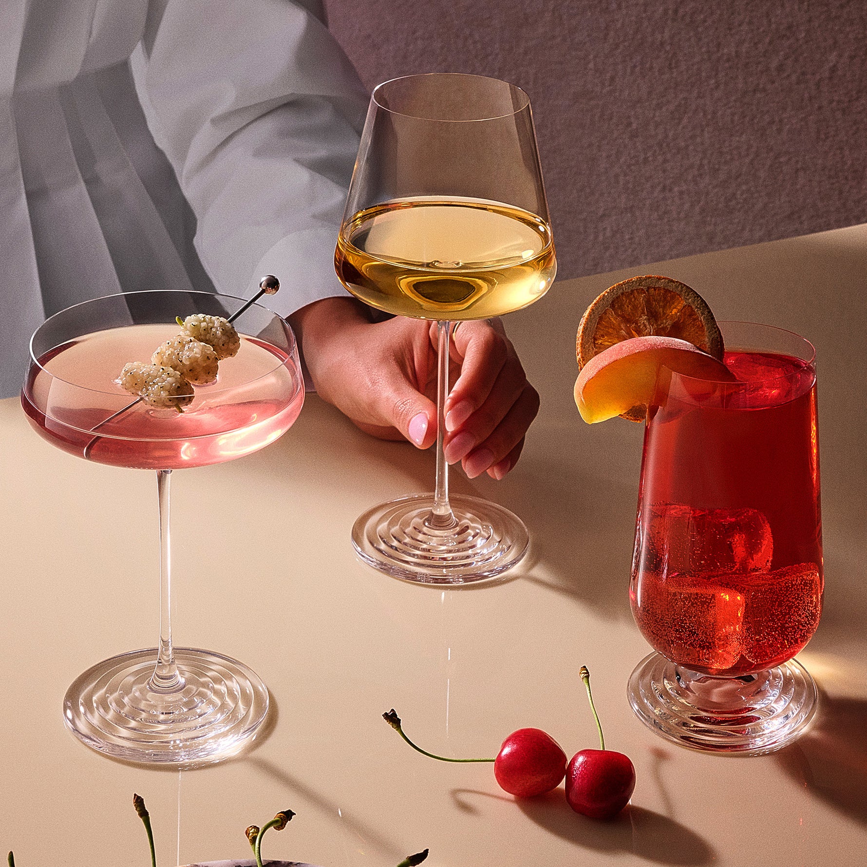 Three clear stemmed drinking glasses of the NUDE x Tom Dixon Splash collection arranged on a beige tabletop with drinks and a person's hand and torso partially visible behind them. The left glass holds a pink cocktail with a cherry garnish, the center glass holds a pale yellow wine, and the right glass holds a red cocktail with a citrus slice and cherry garnish.