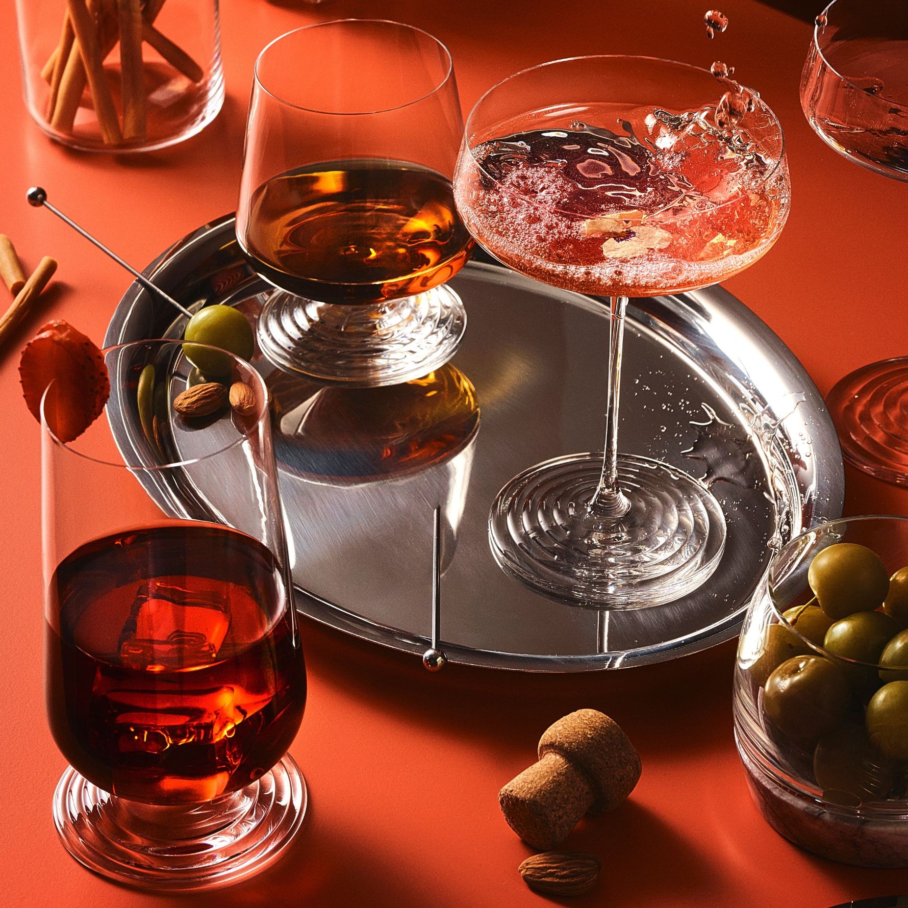 A styled still life of mixed drinks in clear glassware on a reflective silver tray, with olives, corks, and cinnamon sticks on a warm orange-red surface.