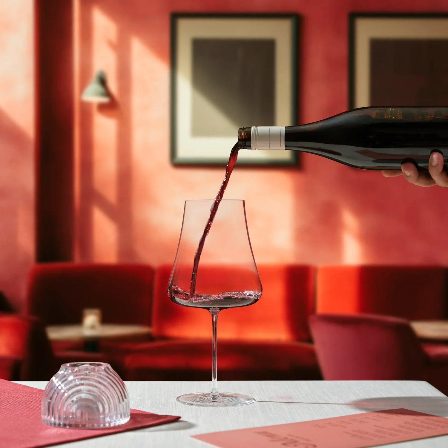 Red wine being poured into a stem zero volcano glass with a blurred restaurant interior in the background.