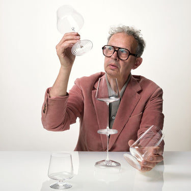 Tom Dixon with gray hair and glasses, wearing a dusty rose corduroy blazer over a gray shirt, holds and examines several clear glass drinking glasses of the NUDE Splash Collection against a clean white background.