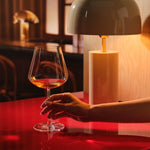 A hand holds a Sera Burgundy glass of orange wine on a glossy red table, lit by a warm table lamp in a dark wood bar setting.