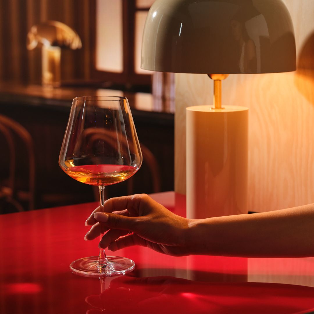 A hand holds a Sera Burgundy glass of orange wine on a glossy red table, lit by a warm table lamp in a dark wood bar setting.