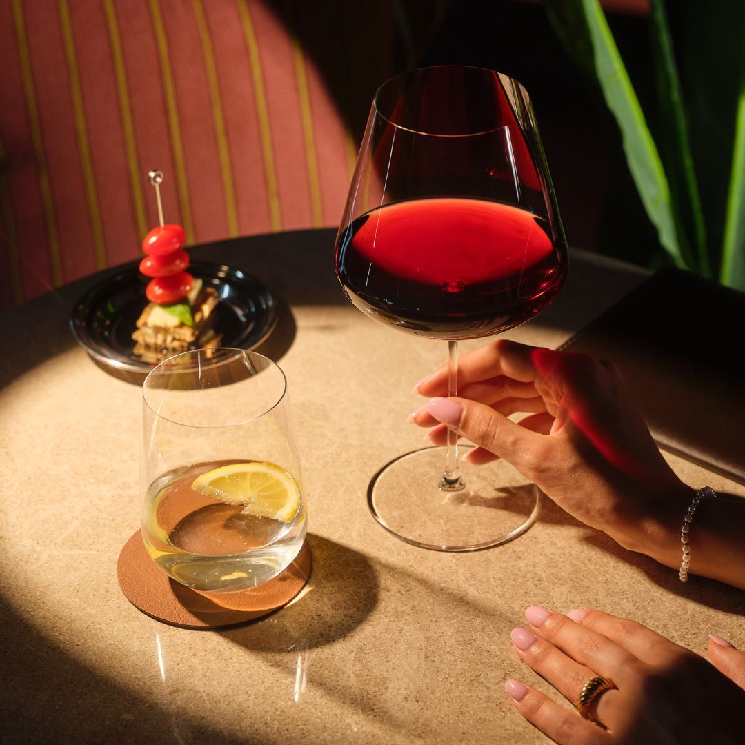 A lifestyle scene on a table showing the Burgundy Sera wine glass partially filled with red wine, alongside the Sera tumbler glass with a drink and garnish. A person's hands are visible holding and resting near the glasses in warm, dramatic lighting.