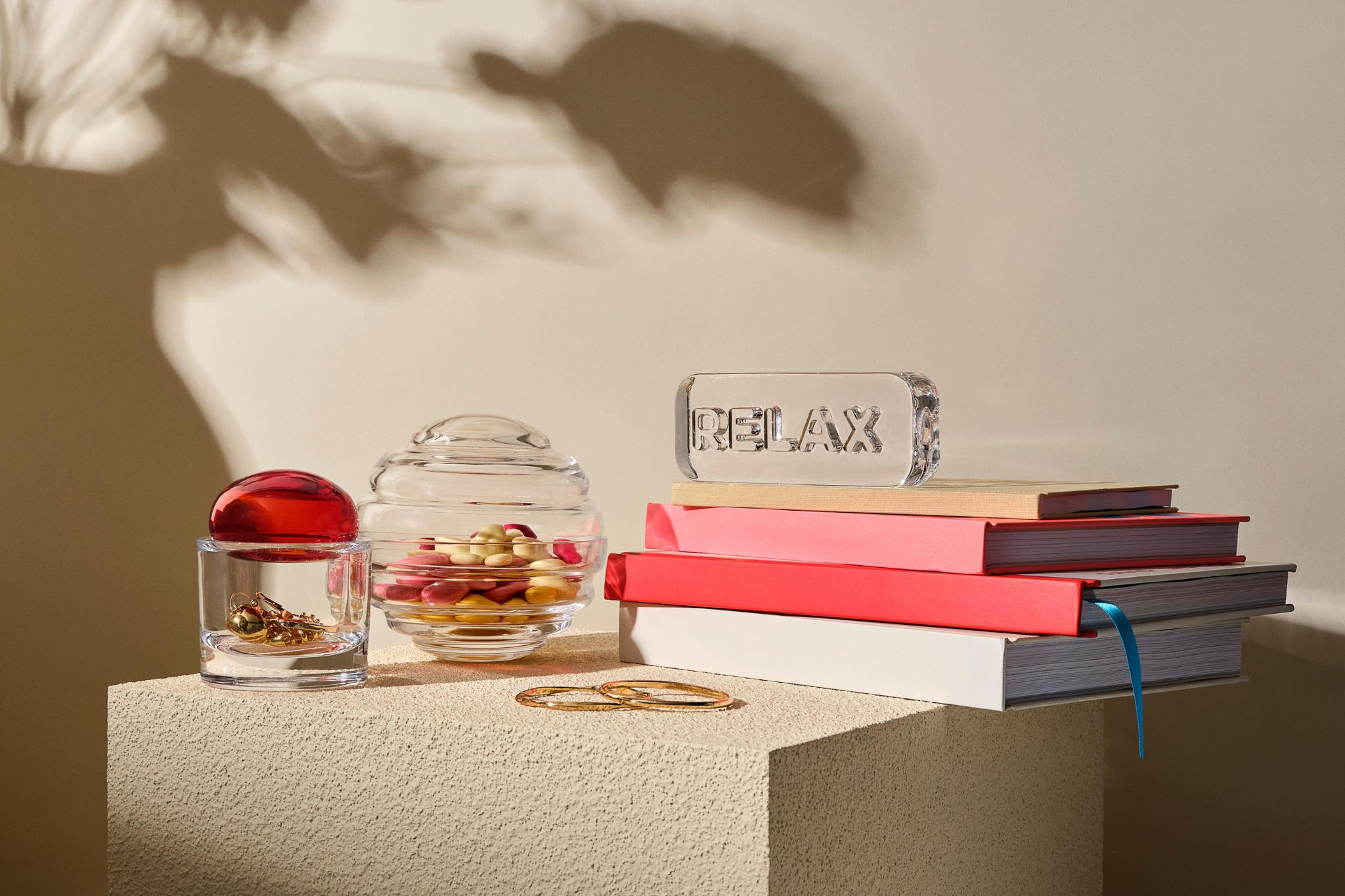 A styled still life on a beige pedestal with a  Nest Candy box a small red-lidded Ecrin container, gold rings, and a stack of books including one with a clear Paroles paper weight reading 'RELAX'.