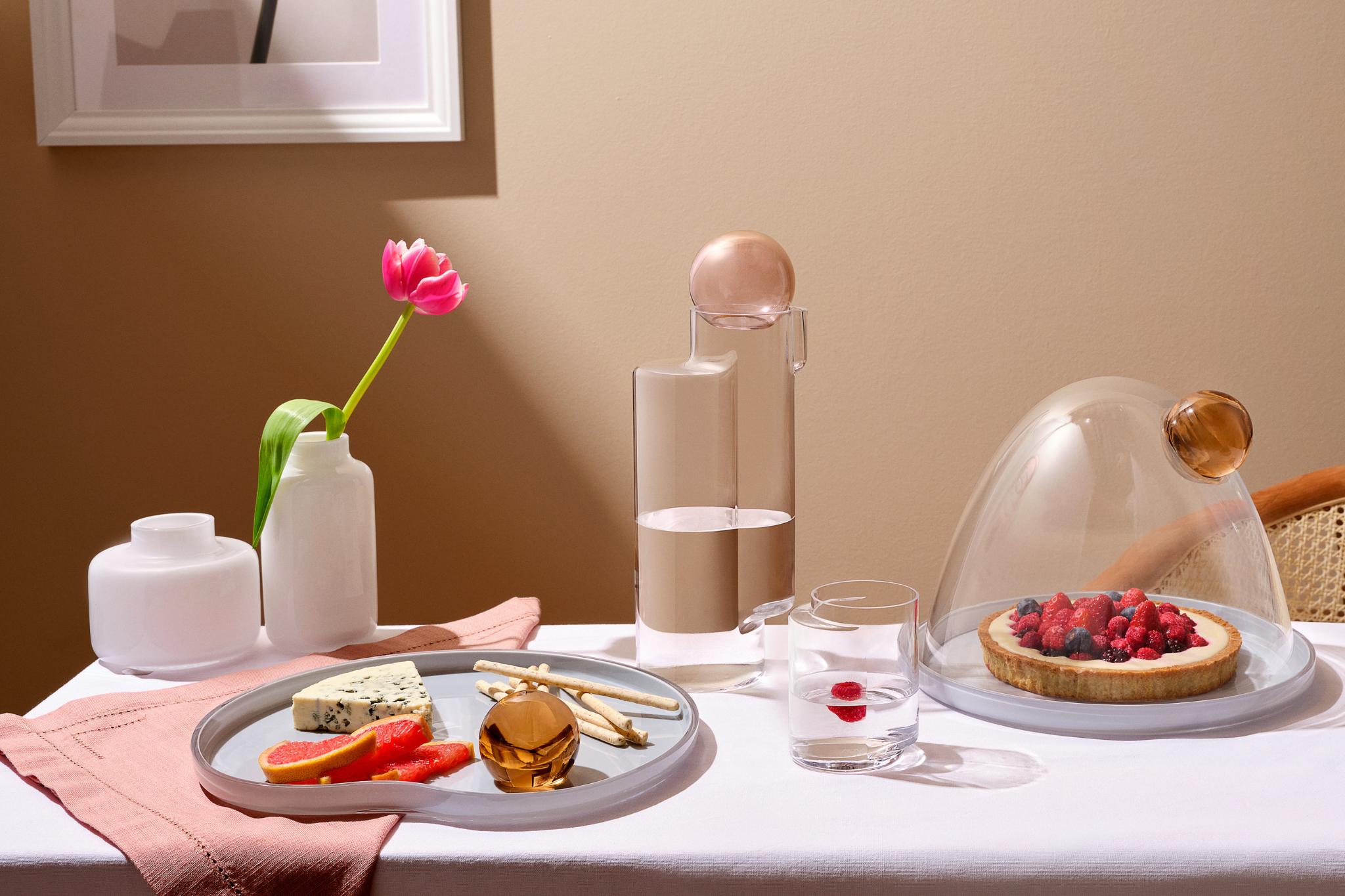 A styled tabletop scene with a white tablecloth, beige wall, a pink tulip in a white Magnolia vase, a Juno water glass  with a cherry, a Juno serving plate with cheese, fruit,  and a tart under a clear Juno cake dome. No person or animal is present, but this is not a clean isolated product shot; it reads as a real interior setting, so Lifestyle fits better than Product/Packaging.