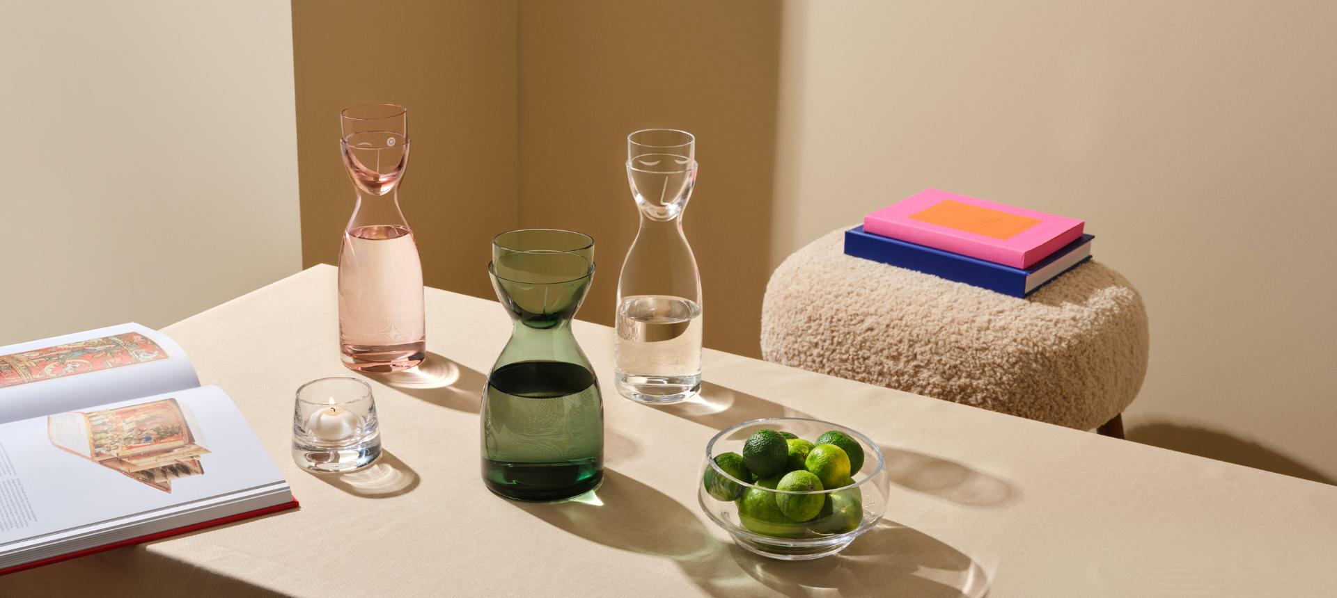 NUDE Mr & Mrs Water Set carafes in pink and green with matching tumblers, Heads Up bowl filled with limes, and Mist votive holder styled on a table with books and a boucle stool.