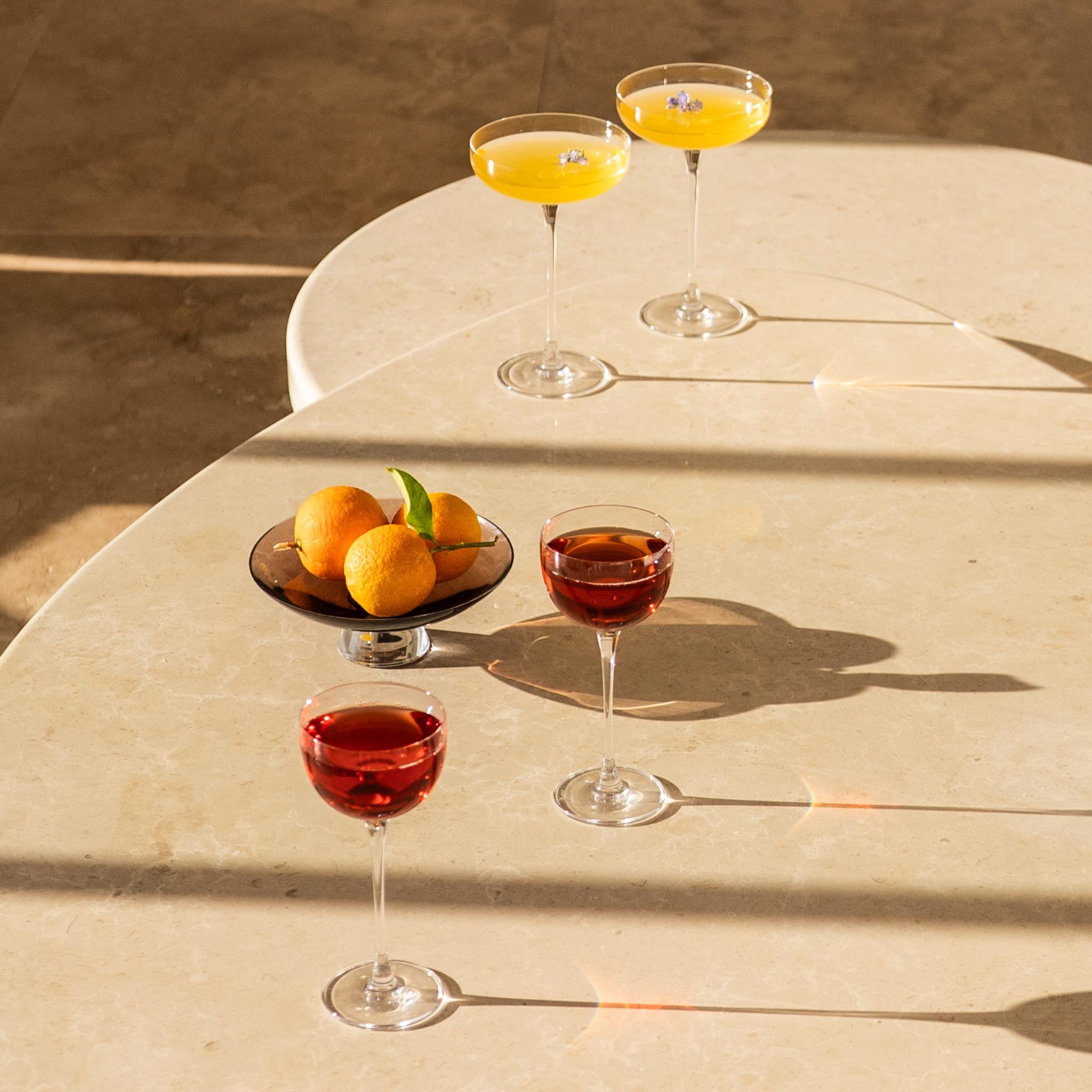 NUDE Savage Remy Coupe and Pony glasses with orange and red cocktails, styled on a marble table with fresh oranges."