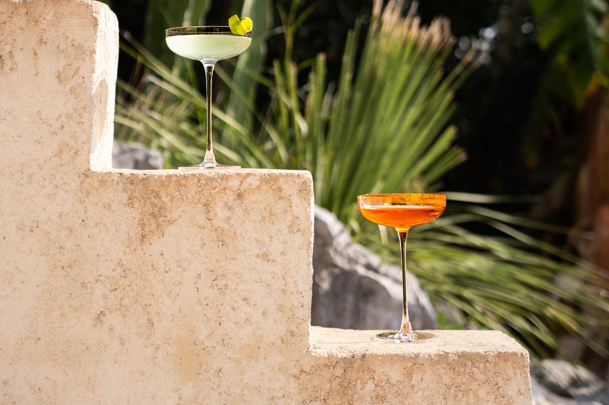 NUDE Savage Remy Coupe cocktail glasses, one with green cocktail and citrus twist, one with orange cocktail, on textured stone steps against greenery.