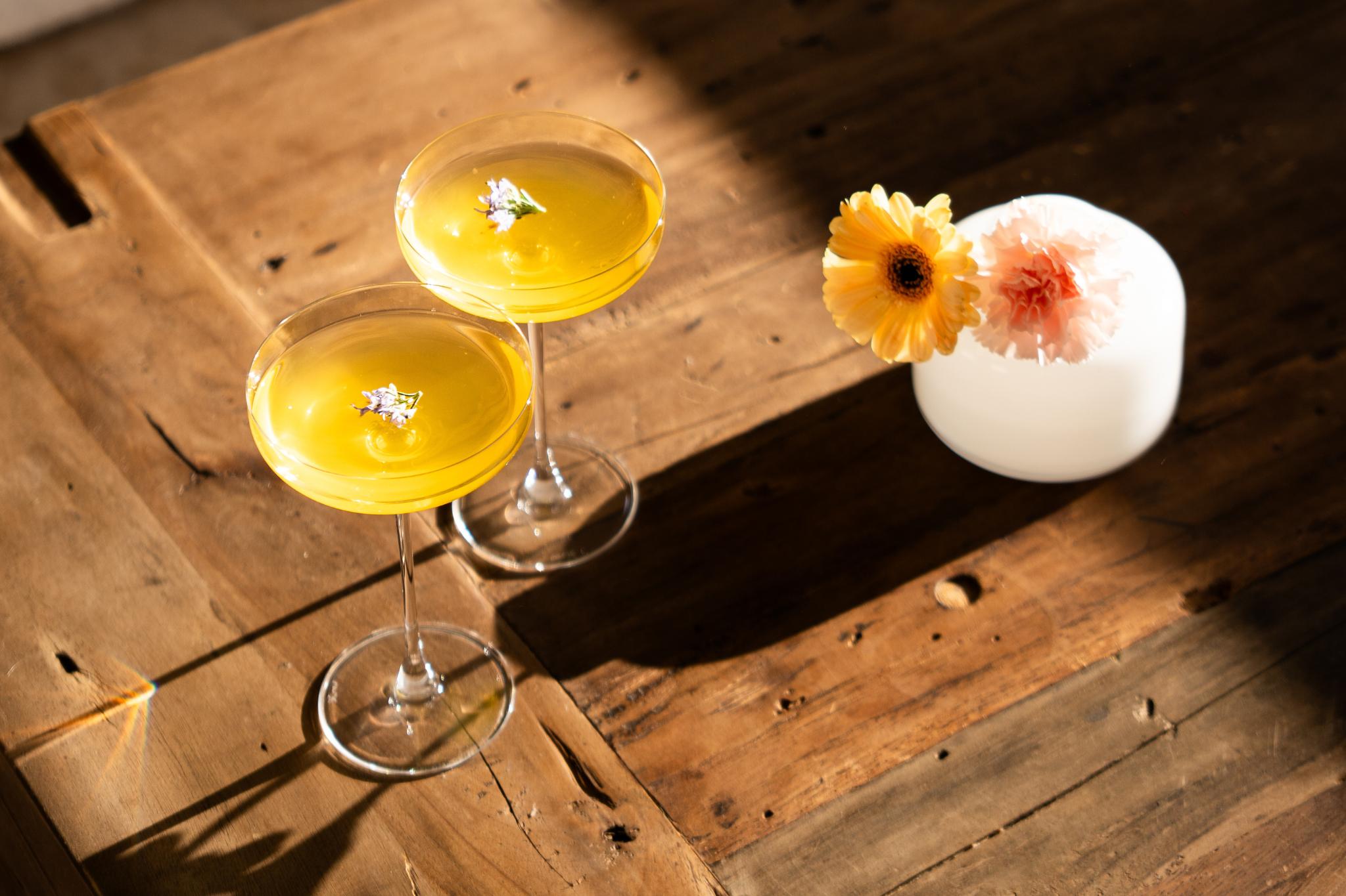 NUDE Savage Remy Coupe cocktail glasses filled with a golden cocktail, garnished with edible flowers, on a rustic wooden table beside fresh flowers in a white Magnolia vase.