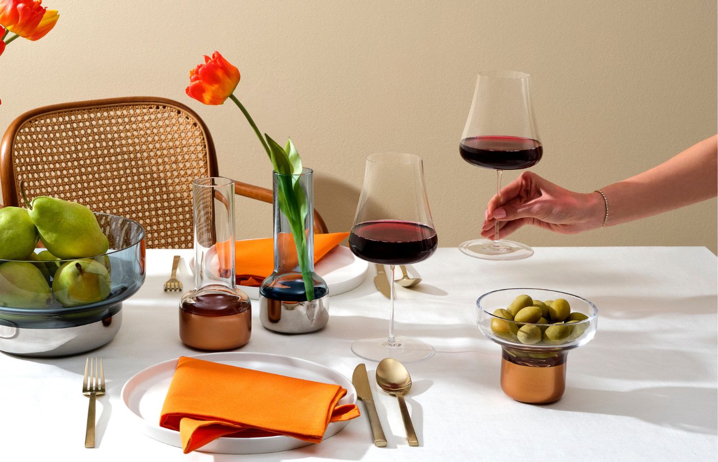 NUDE Stem Zero Volcano Red Wine Glass held by hand at a styled table setting with copper-based vases, decanter, and fresh fruit.