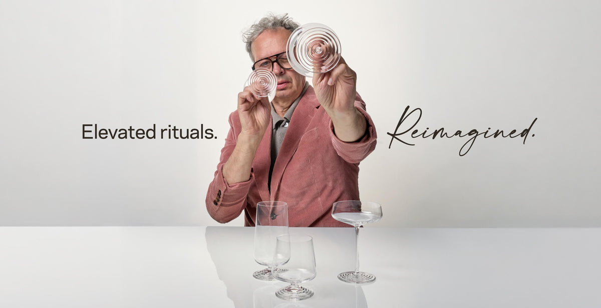 Tom Dixon in a pink corduroy jacket holding two NUDE Splash glasses up to his glasses, with three more Splash glasses on a table below. Text reads: Elevated rituals. Reimagined