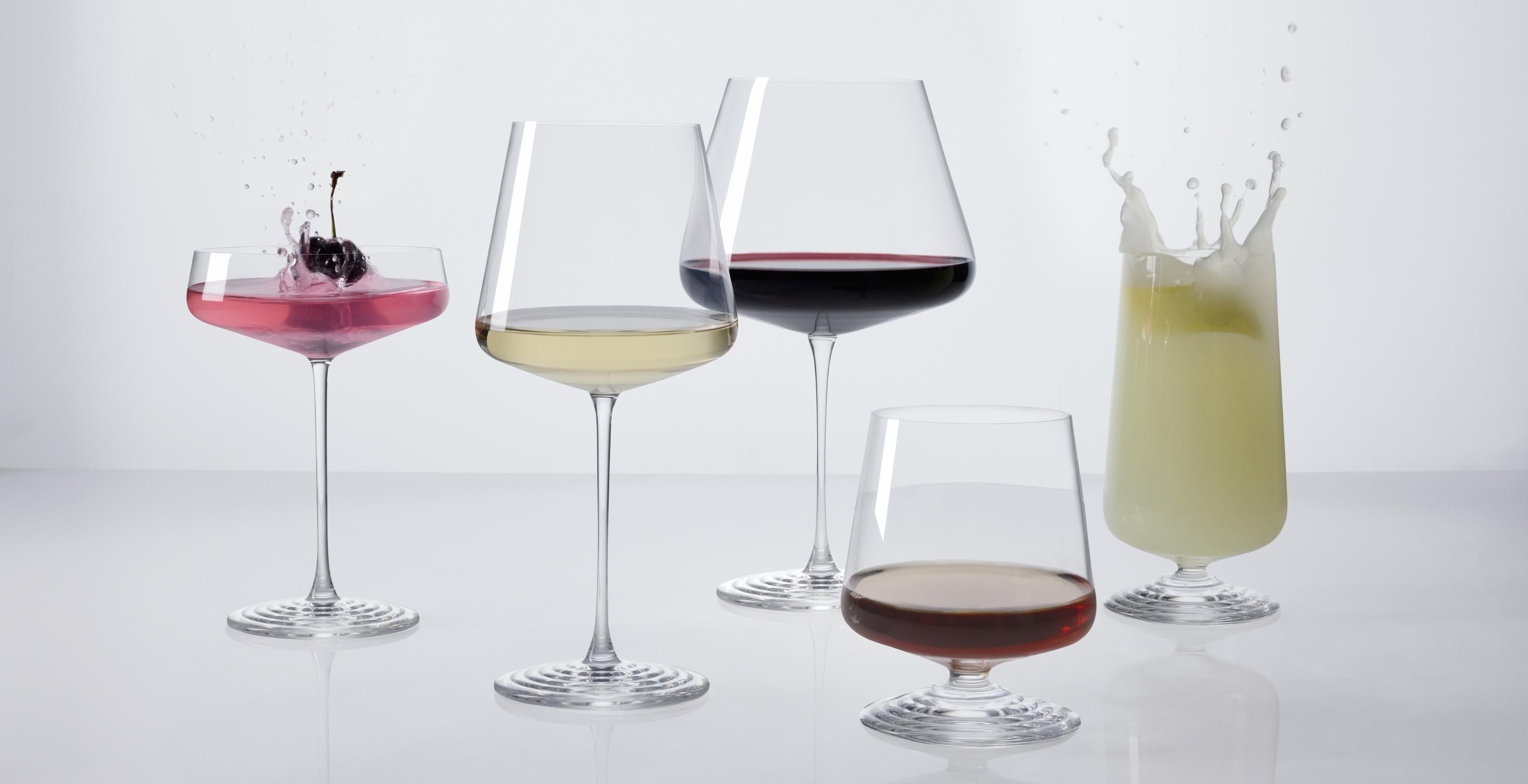 The five NUDE Splash glasses in a row, each filled: coupe with pink cocktail and cherry garnish splashing, white wine, red wine, lowball with amber spirit, and highball with a pale green drink splashing, all on a white reflective surface.
