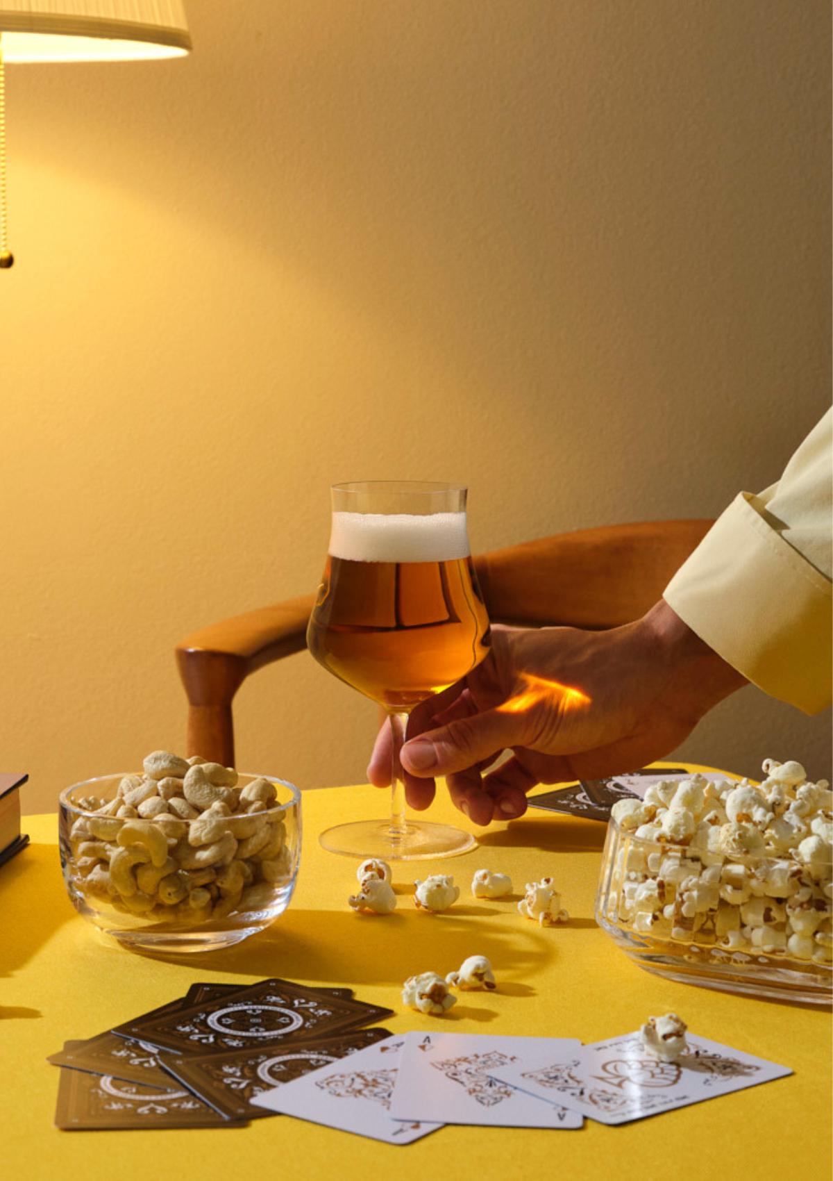 A hand holding a NUDE Islands beer glass of golden beer, with a NUDE Big Top bowl and Malt bowl on a warm, lamplit evening table.