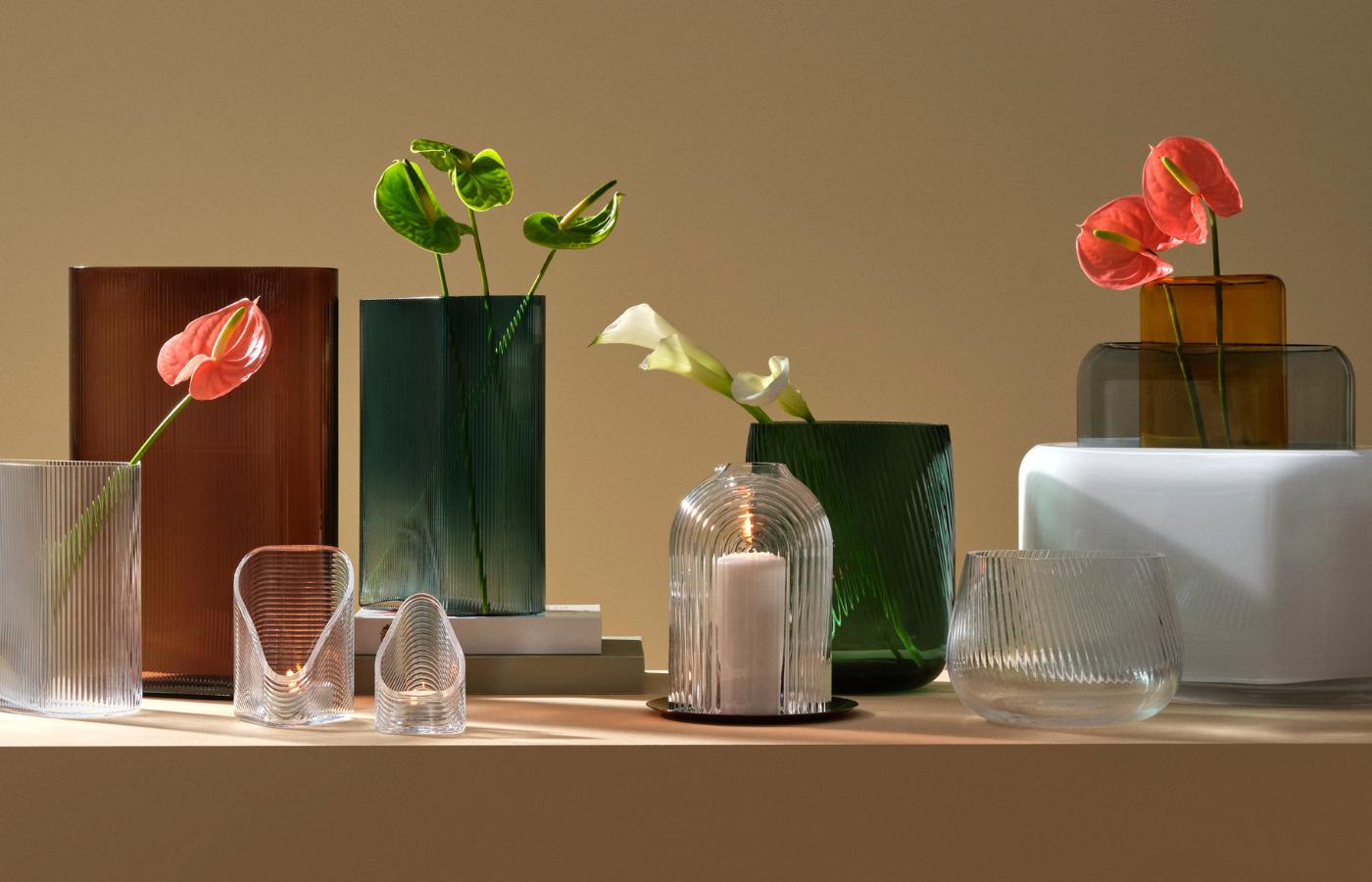 A grouping of amber, petrol, clear and white NUDE Mist, Opti and Layers vases with an Ilo candleholder, styled with flowers on a sunlit shelf.