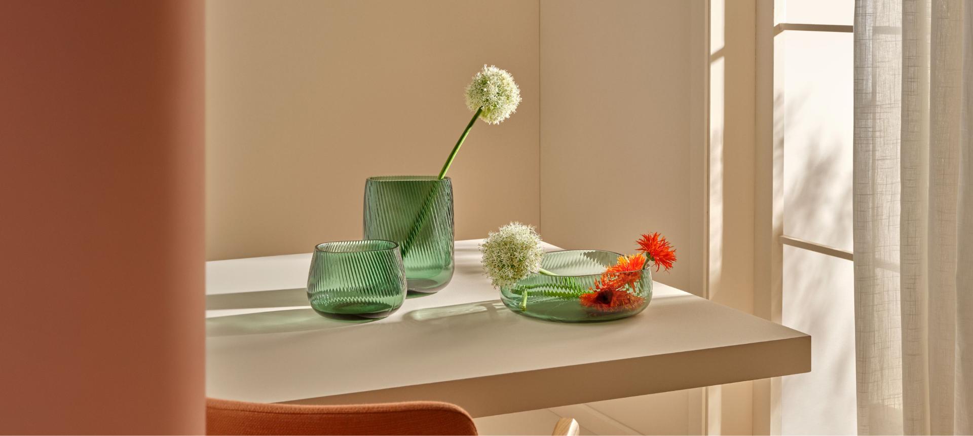 A tall and medium green NUDE Opti vase and Opti centre piece bowl with flowers on a sunlit table.