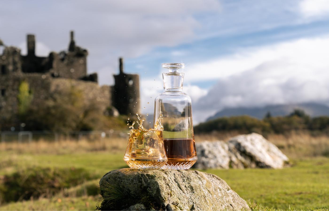 NUDE Paris whisky decanter and glass with whisky splash on a mossy rock in front of a ruined Scottish castle, photography by Murray Anderson