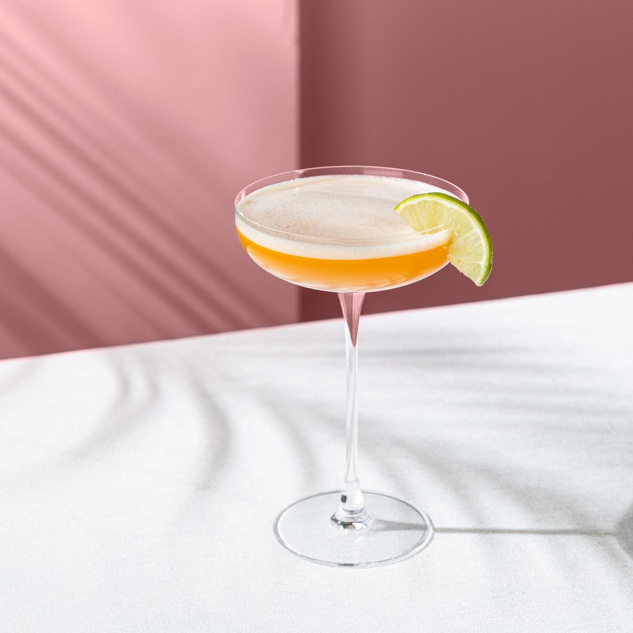 NUDE Savage Coupe Glass with an amber cocktail and lime wedge on a blush backdrop.