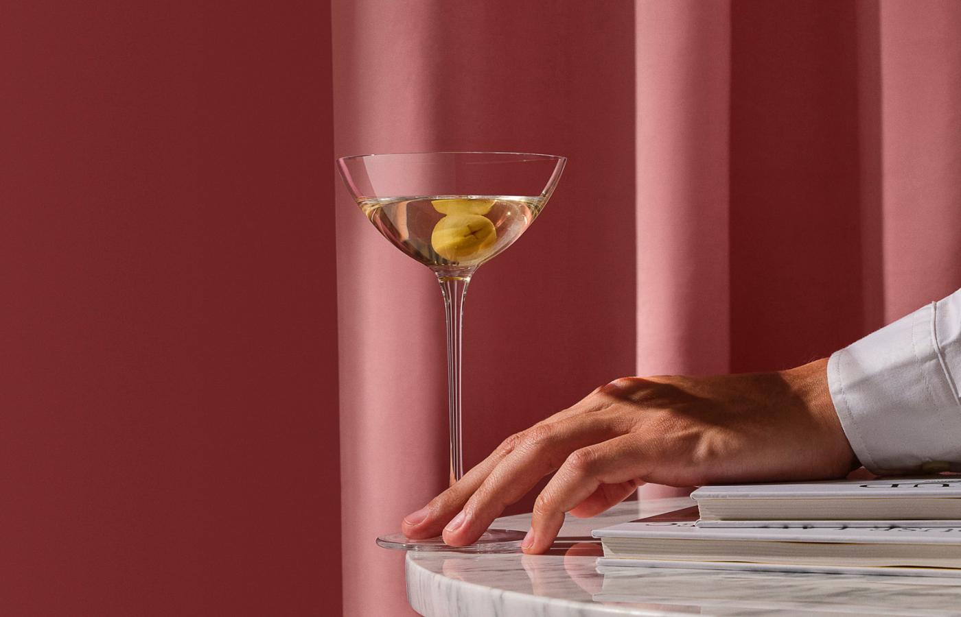 A martini with an olive in a NUDE Savage Coupetini Glass on a marble table.