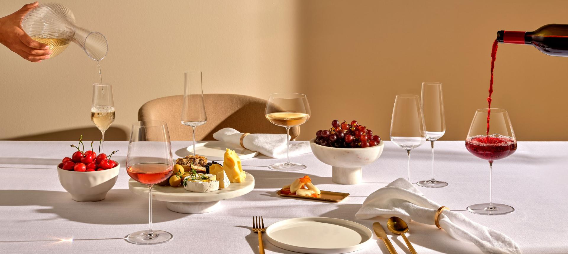 NUDE Stem Zero Full Bodied White Wine Glass, Elegant Medium Volcano Champagne Glass, and Delicate White Flute, red wine being poured at a set table with grapes, cheese, and gold flatware.