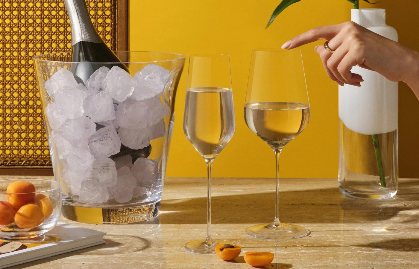 Two wine glasses with ice and champagne, a hand pointing at one glass, on a wooden table with a yellow background.