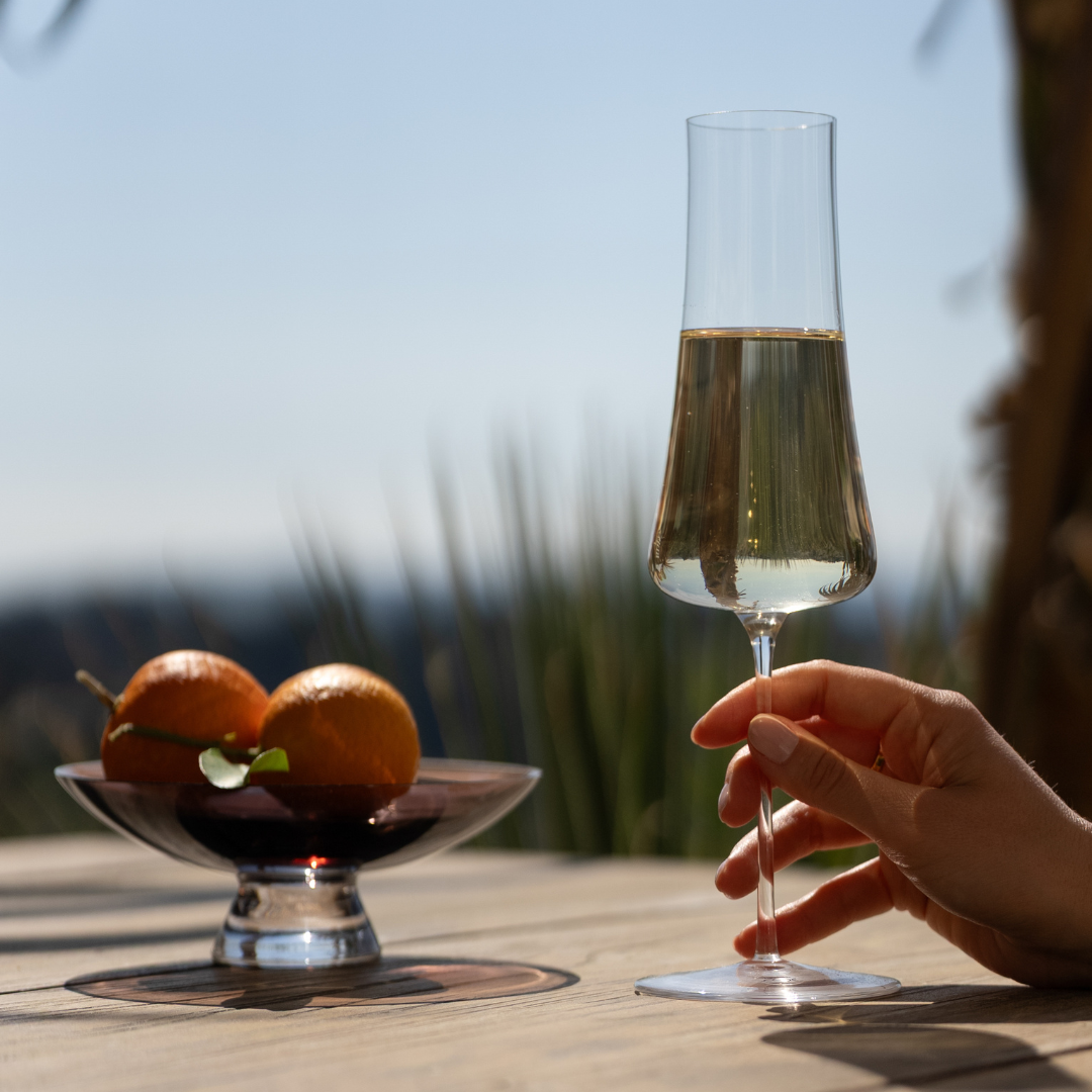 NUDE Stem Zero Volcano champagne glass held by hand on a sunlit outdoor terrace with citrus fruit bowl and open sky.