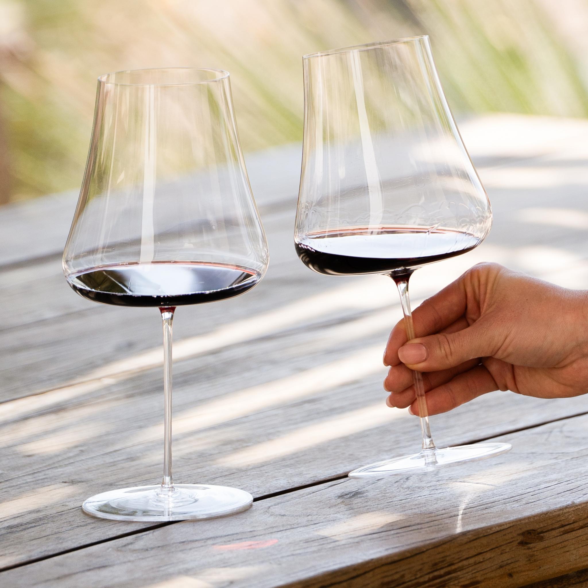 NUDE Stem Zero Volcano Red Wine Glass, two glasses filled with red wine on a wooden outdoor table, one held by hand