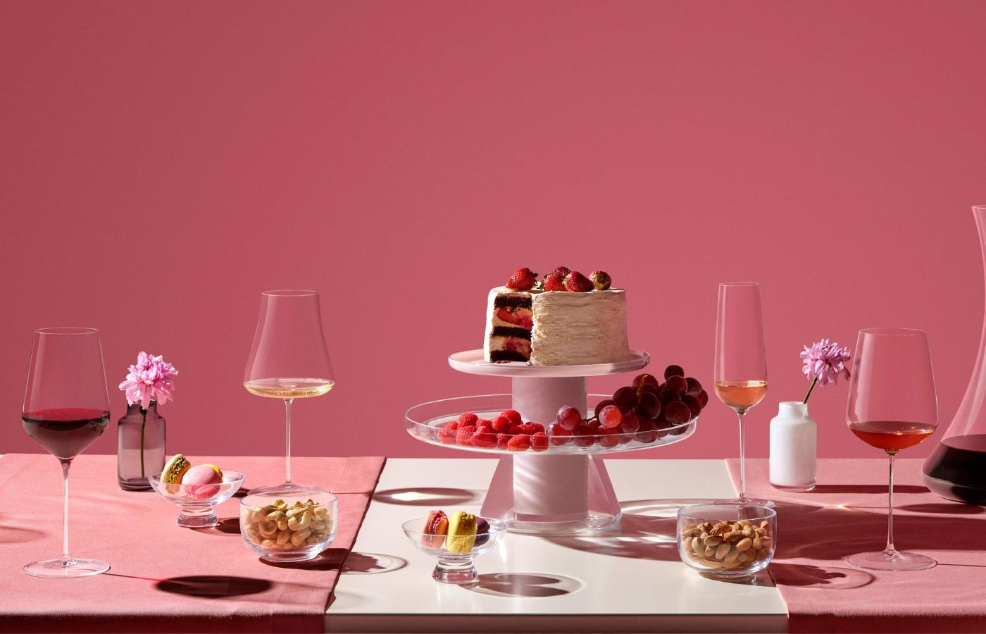 "NUDE Stem Zero Volcano, Trio, and Vertigo glasses, styled dessert table setting with cake, fruit, and macarons against a pink backdrop.
